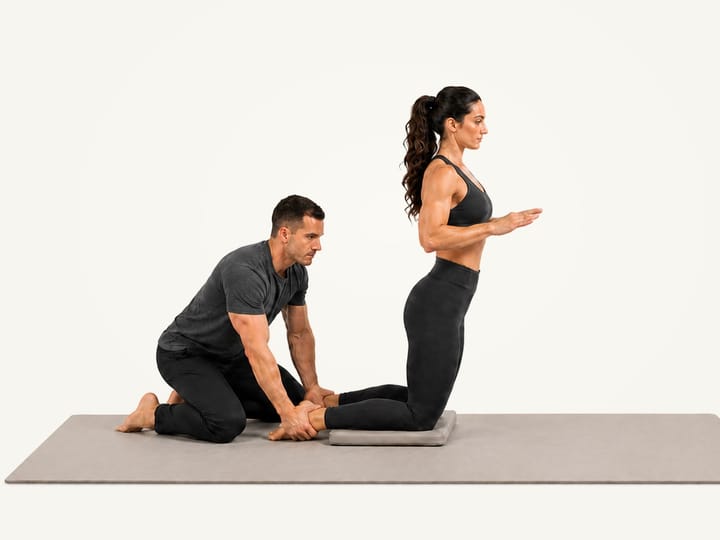 Step 1 of the Nordic curl: kneeling tall on a pad, a partner kneeling behind and holding both ankles